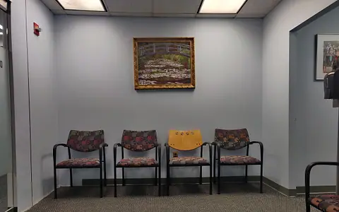 An empty medical waiting room with four chairs against a pale wall, a framed picture and a wall-mounted hand-sanitiser dispenser.
