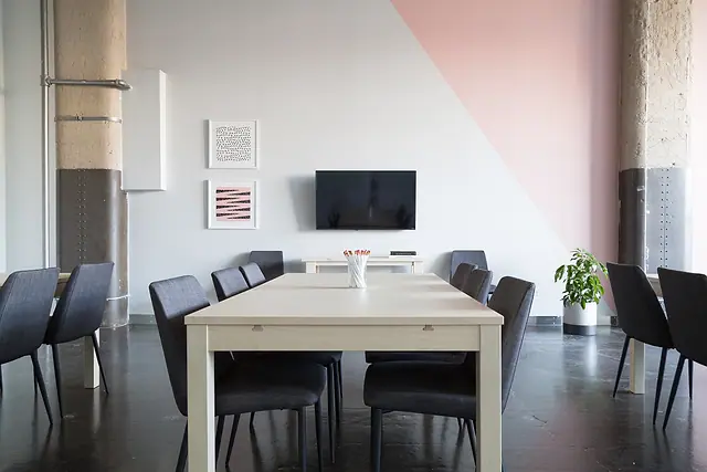 An empty meeting room in a commercial building, with a light wood table, dark chairs and a wall-mounted screen.