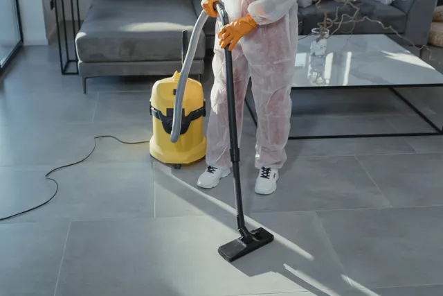 A person in a white protective coverall and gloves vacuuming a tiled floor with a canister vacuum.