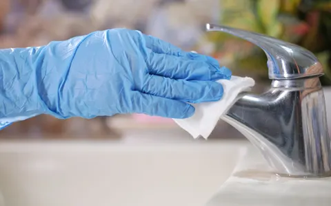 A gloved hand wiping a chrome mixer tap with a white cloth at the edge of a white sink.