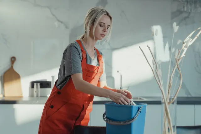 A person in work overalls rinsing a cloth in a bucket beside a kitchen counter.
