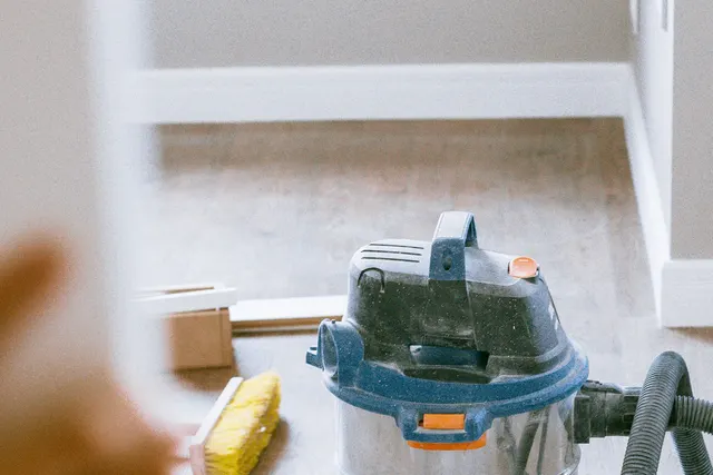 A wet-and-dry vacuum and a broom standing on a dusty wood floor in a room being finished after renovation work.