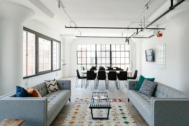 A shared lounge and meeting area in a commercial building, with sofas, a low table and a long meeting table by tall windows.